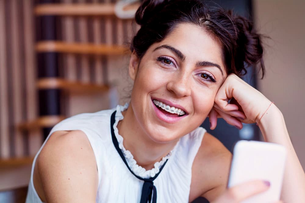Smiling confidently, a young woman with dark hair in buns and braces holds her smartphone indoors—demonstrating the results of adult orthodontic care at Andros Orthodontics in Pasco, WA.