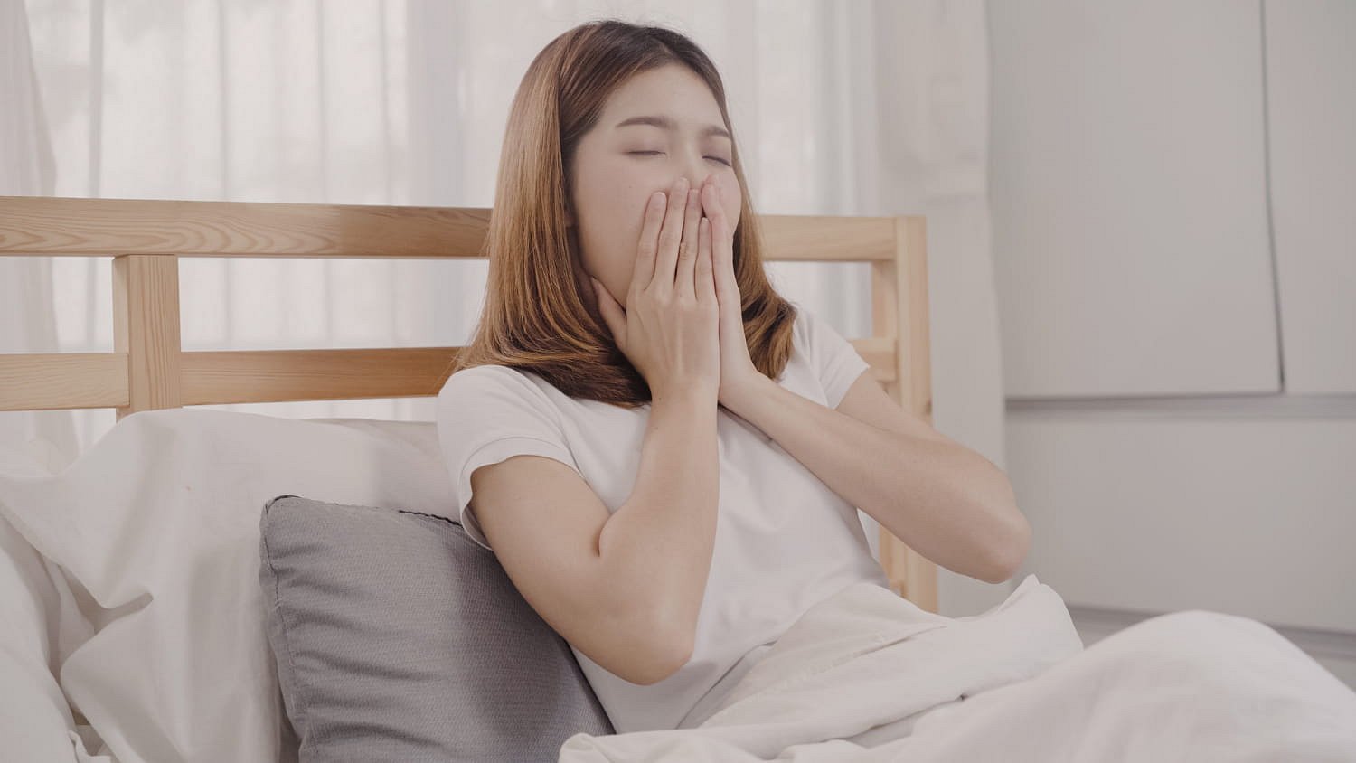 A woman in bed covers her mouth while yawning, showing signs of habitual mouth breathing—a concern Andros Orthodontics can address in Pasco, WA.