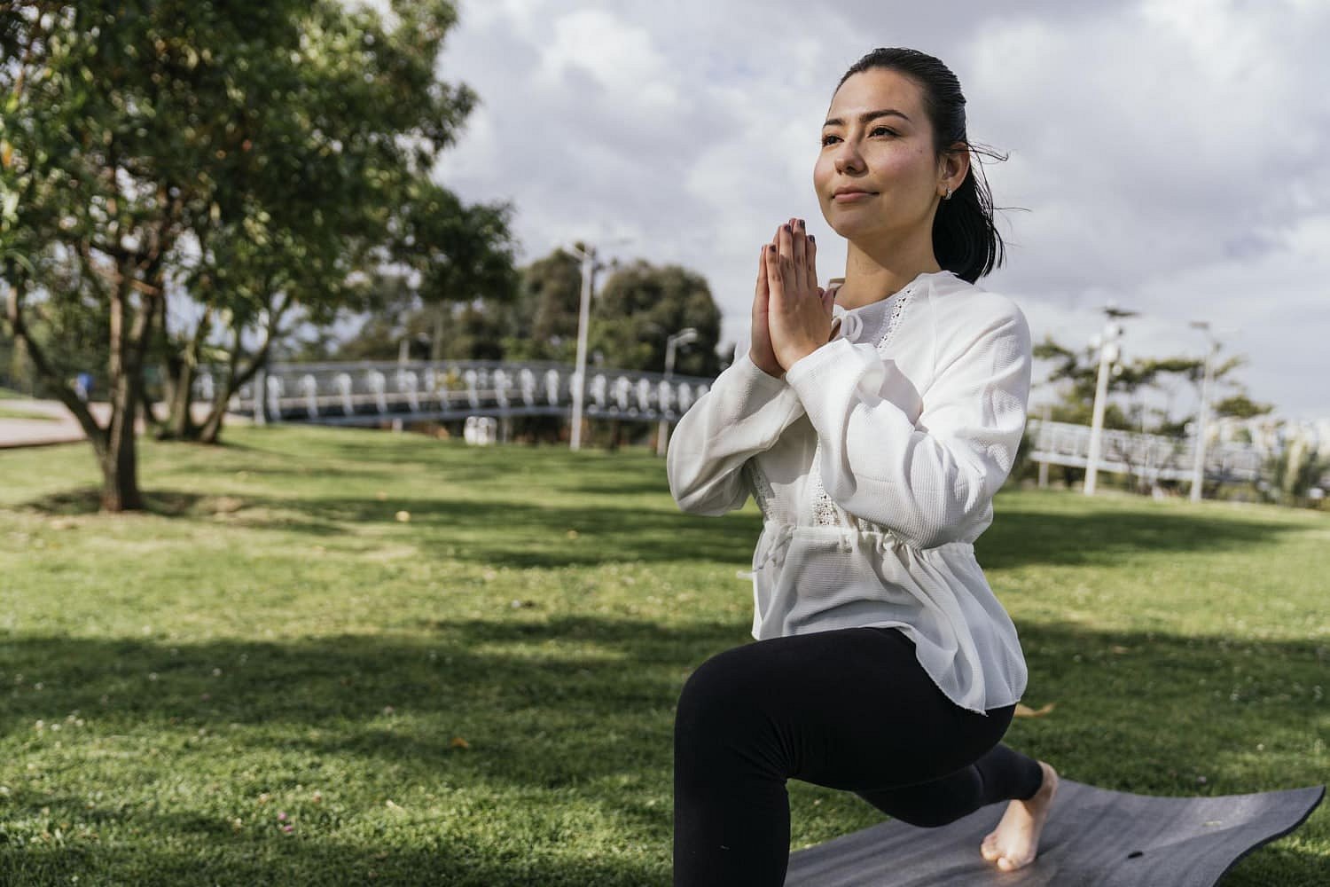 Practicing yoga in a grassy park, a woman holds a lunge pose with hands together and focuses on mindful breathing—not just mouth breathing. Andros Orthodontics in Pasco, WA encourages healthy habits like this for overall wellness.
