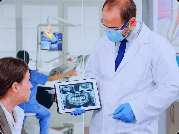 At Andros Orthodontics in Pasco, WA, a dentist wearing a mask and gloves explains jaw surgery options to a seated female patient by showing her a dental X-ray on a tablet in their modern clinic.