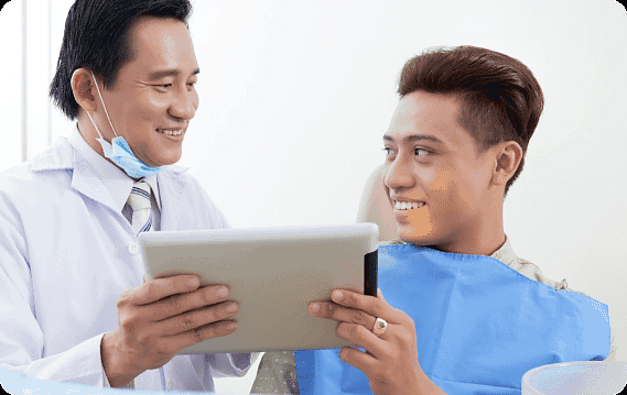 At Andros Orthodontics in Pasco, WA, a dentist reviews jaw surgery options with a smiling patient in the dental chair as they both focus on information displayed on a tablet. The patient is wearing a blue dental bib.