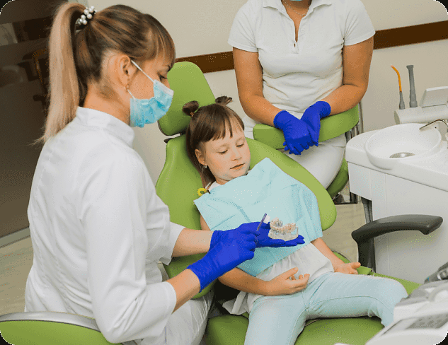 At Andros Orthodontics in Pasco, WA, a female children's orthodontist shows a dental model to a young girl in the chair, while another dental professional looks on. Both staff members are dressed in white uniforms and blue gloves.