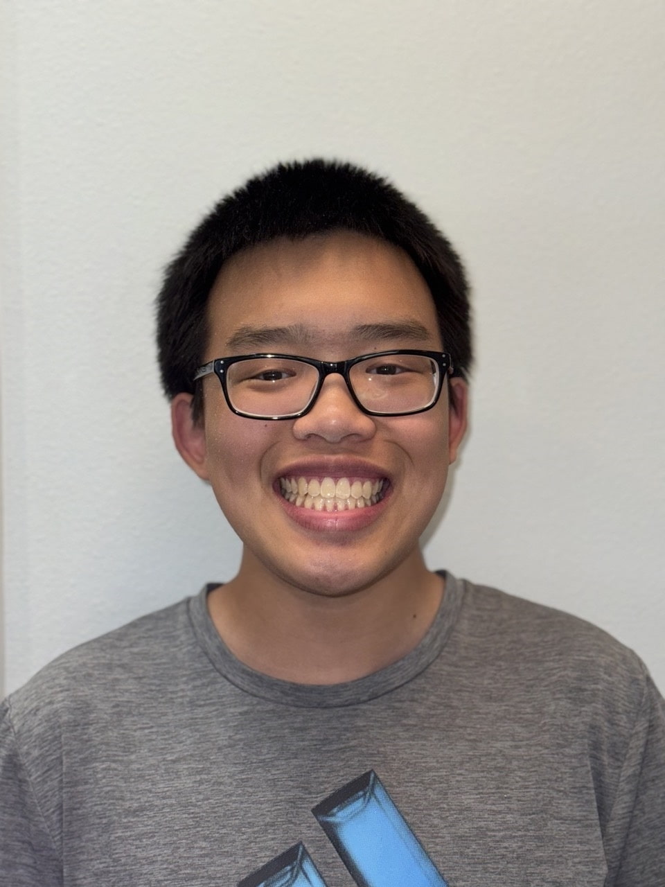 A young person with short black hair and glasses confidently shows off their post-braces smile in a gray T-shirt with a blue design, standing against a plain light-colored wall at Andros Orthodontics in Pasco, WA.