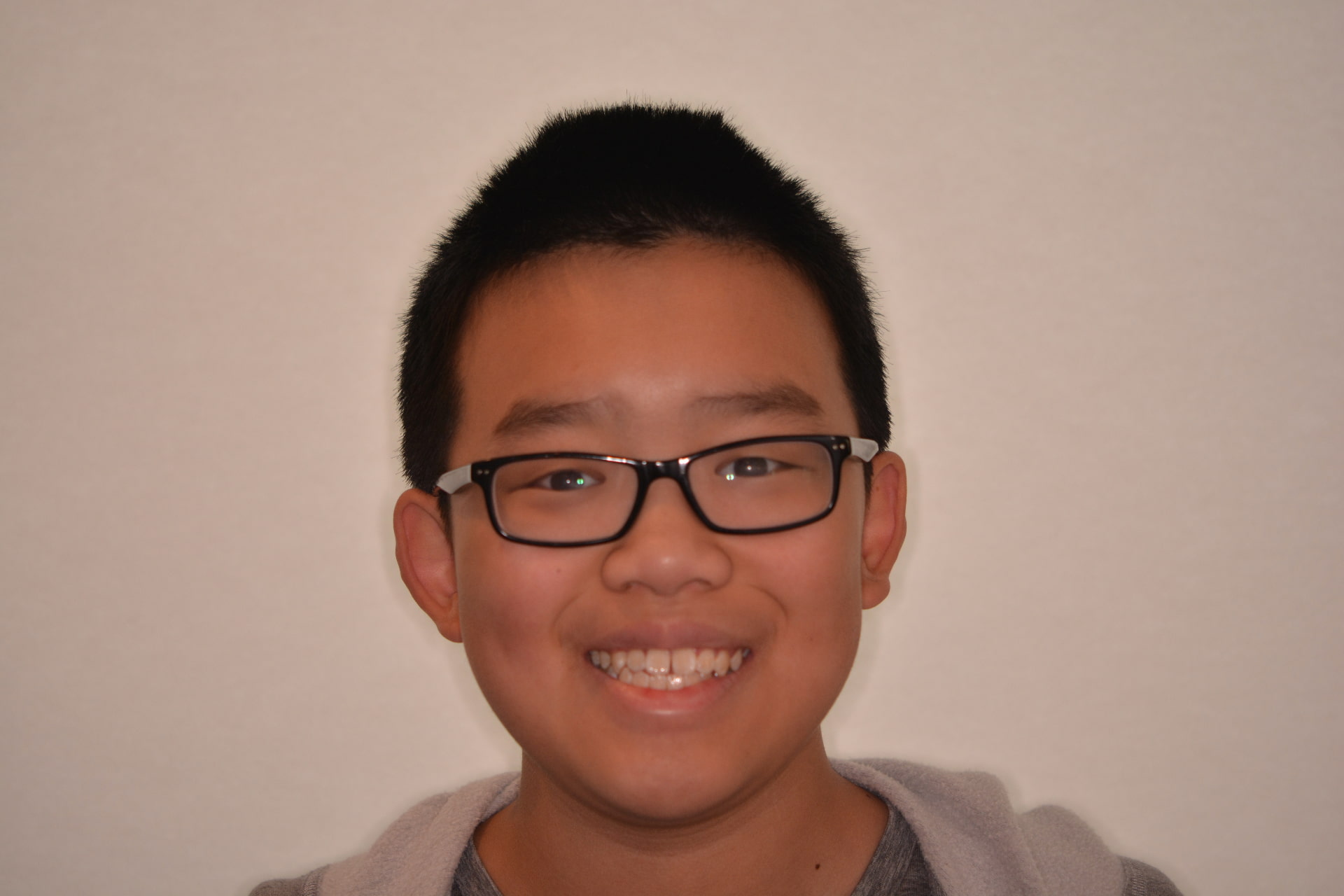 A young person with short black hair and glasses shows their natural teeth before braces, smiling for Andros Orthodontics in Pasco, WA against a plain light background.