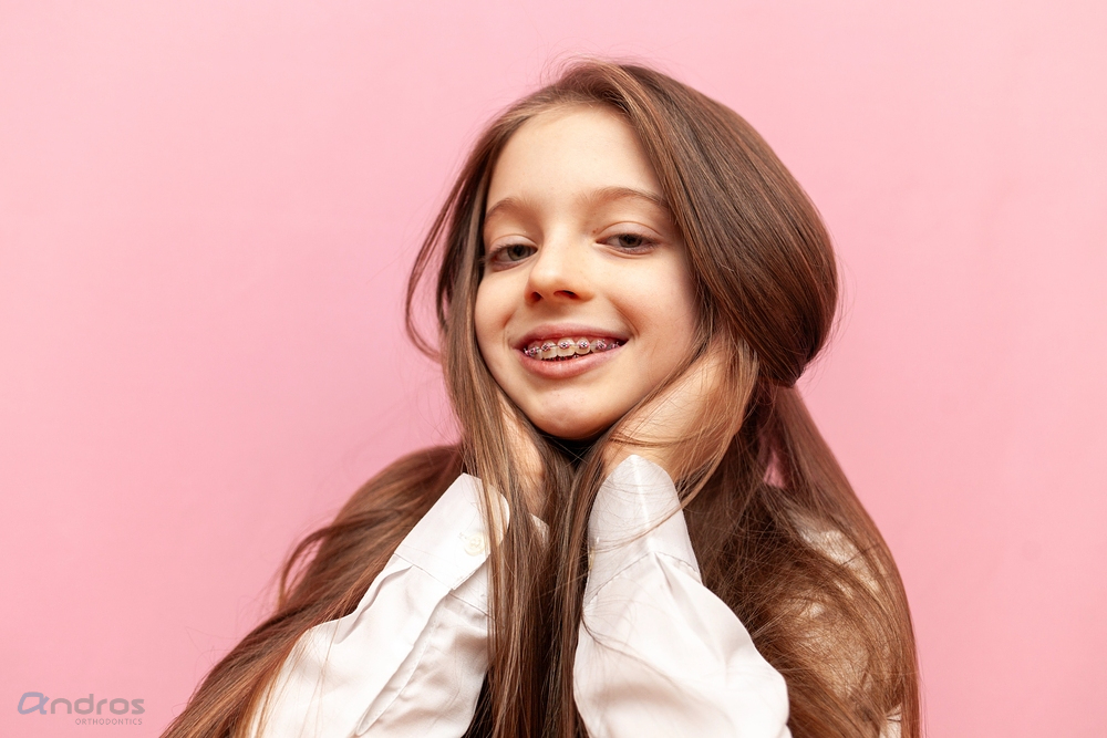 A young girl with long brown hair and braces, treated at Andros Orthodontics' braces for overbite in Pasco, WA, smiles with her hands on her cheeks against a pink background.