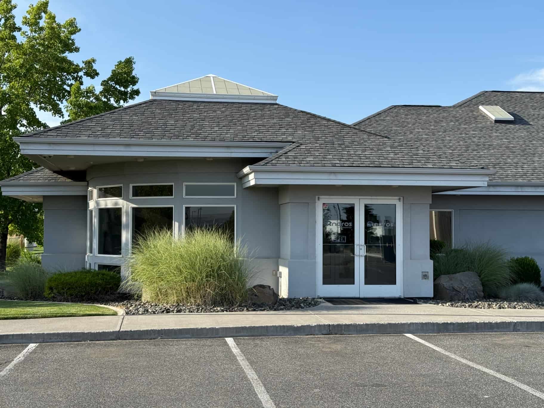Andros Orthodontics in Pasco, WA, is located in a single-story office building featuring gray walls, a dark shingle roof, white trim, and glass double doors. The building is surrounded by shrubs and grasses and has an empty parking lot in front.