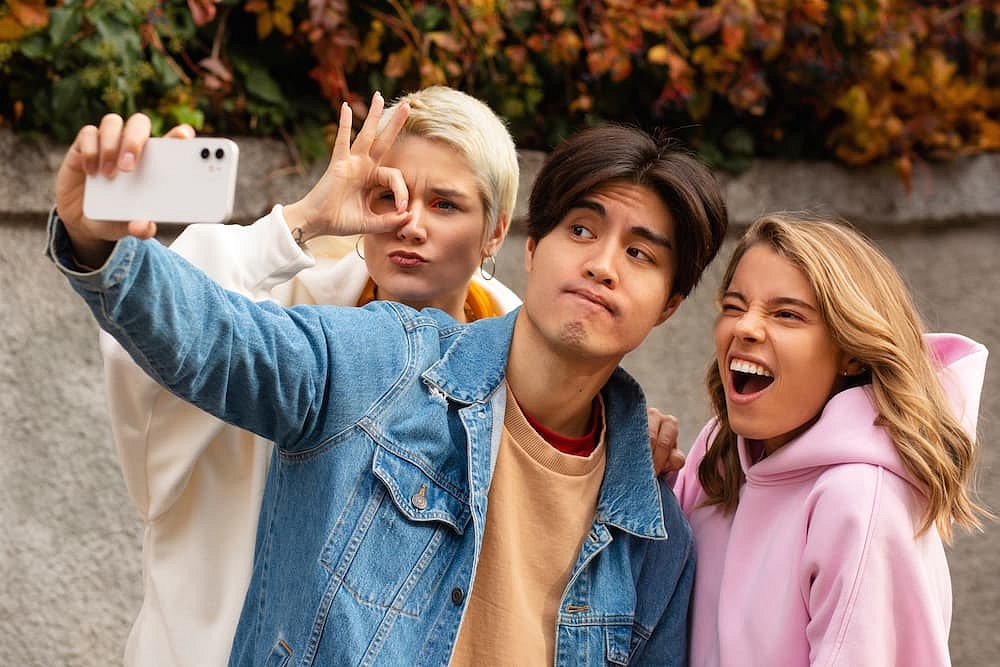 Three young adults take a selfie among autumn leaves—one flashes a hand sign, another reveals their braces from Andros Orthodontics in Pasco, WA with a wide smile, showcasing confidence from different Types of Braces, while the third gazes neutrally at the camera.