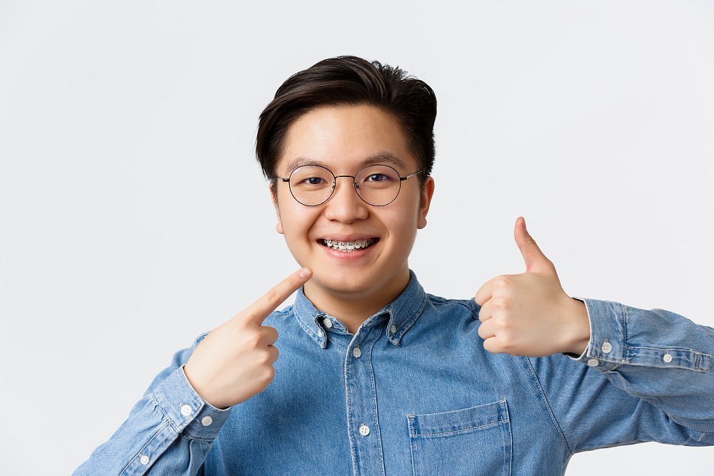 A young person in glasses and a denim shirt smiles confidently, pointing to their adult braces and giving a thumbs-up—showcasing their Andros Orthodontics results in Pasco, WA.