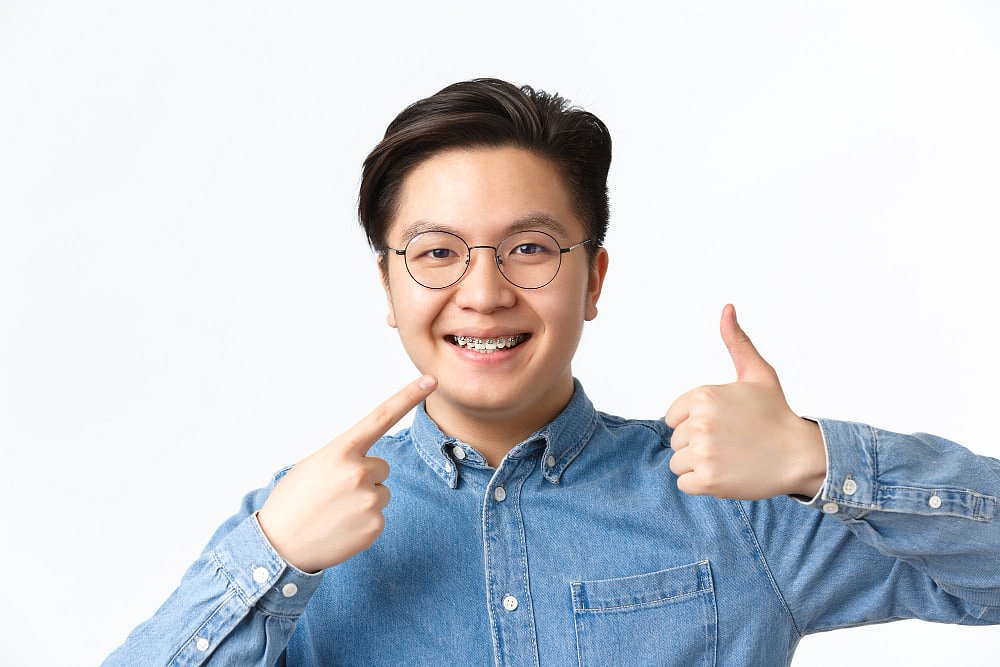At Andros Orthodontics in Pasco, WA, a young person wearing glasses and a denim shirt confidently smiles with braces, pointing to their teeth and giving a thumbs-up—demonstrating the benefits of Adult Orthodontics and Metal Braces.