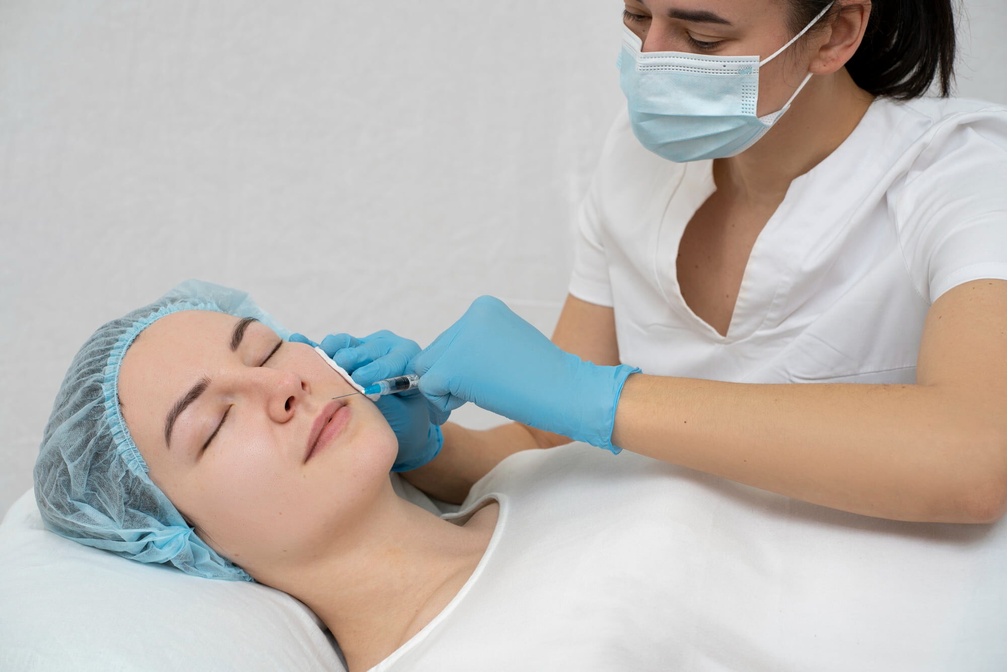 At Andros Orthodontics in Pasco WA, a medical professional in gloves and a mask administers an injection to a woman's cheek as she lies with her eyes closed, dressed in a hair cap and white gown.