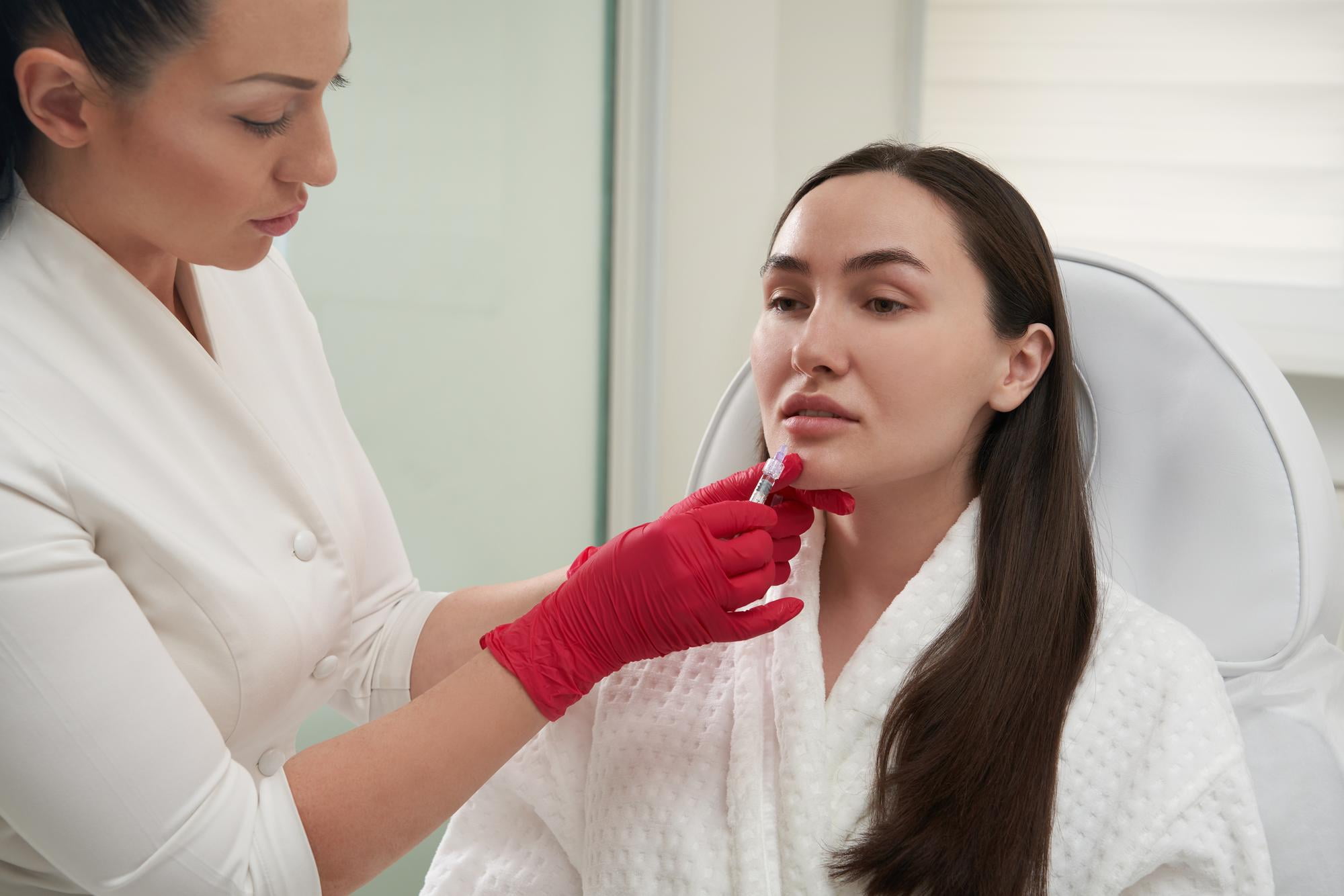At Andros Orthodontics in Pasco WA, a professional in red gloves administers a chin injection to a woman in a white robe, illustrating how Botox can help treat TMJ in a clinical environment.