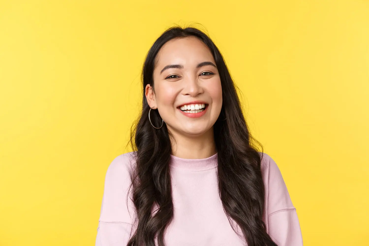 A woman with long dark hair, hoop earrings, and a light pink sweater smiles in front of a yellow background, embodying the welcoming atmosphere at Andros Orthodontics in Pasco, WA.