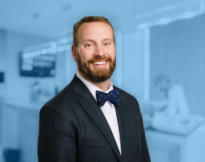 Dr. Nicholas Andros is pictured smiling at the camera in a dark suit with a white shirt and navy blue bow tie against a plain gray background. Dr. Nicholas Andros is orthodontics expert at Andros Orthodontics in Pasco, WA