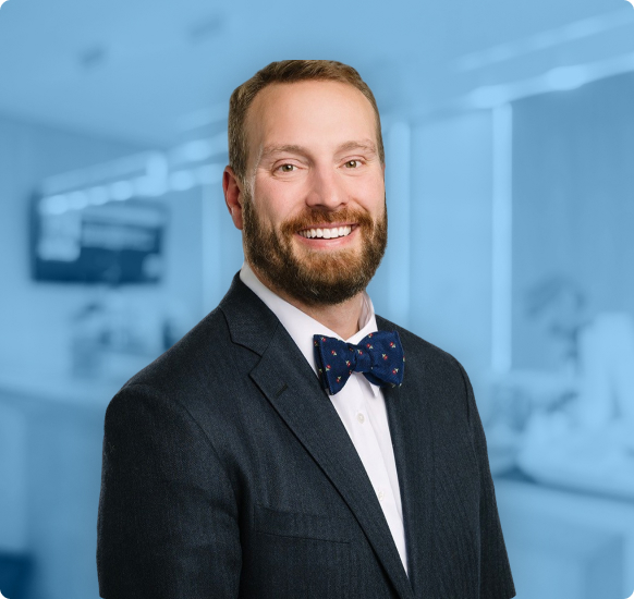Dr. Nicholas Andros is pictured smiling at the camera in a dark suit with a white shirt and navy blue bow tie against a plain gray background. Dr. Nicholas Andros is orthodontics expert at Andros Orthodontics in Pasco, WA