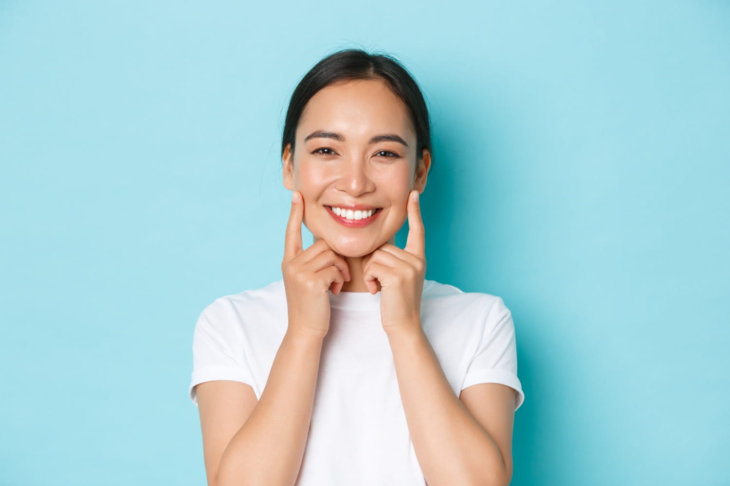 Showing off her confident smile after a visit to Andros Orthodontics in Pasco, WA, a woman in a white t-shirt smiles and points to her cheeks against a blue background.