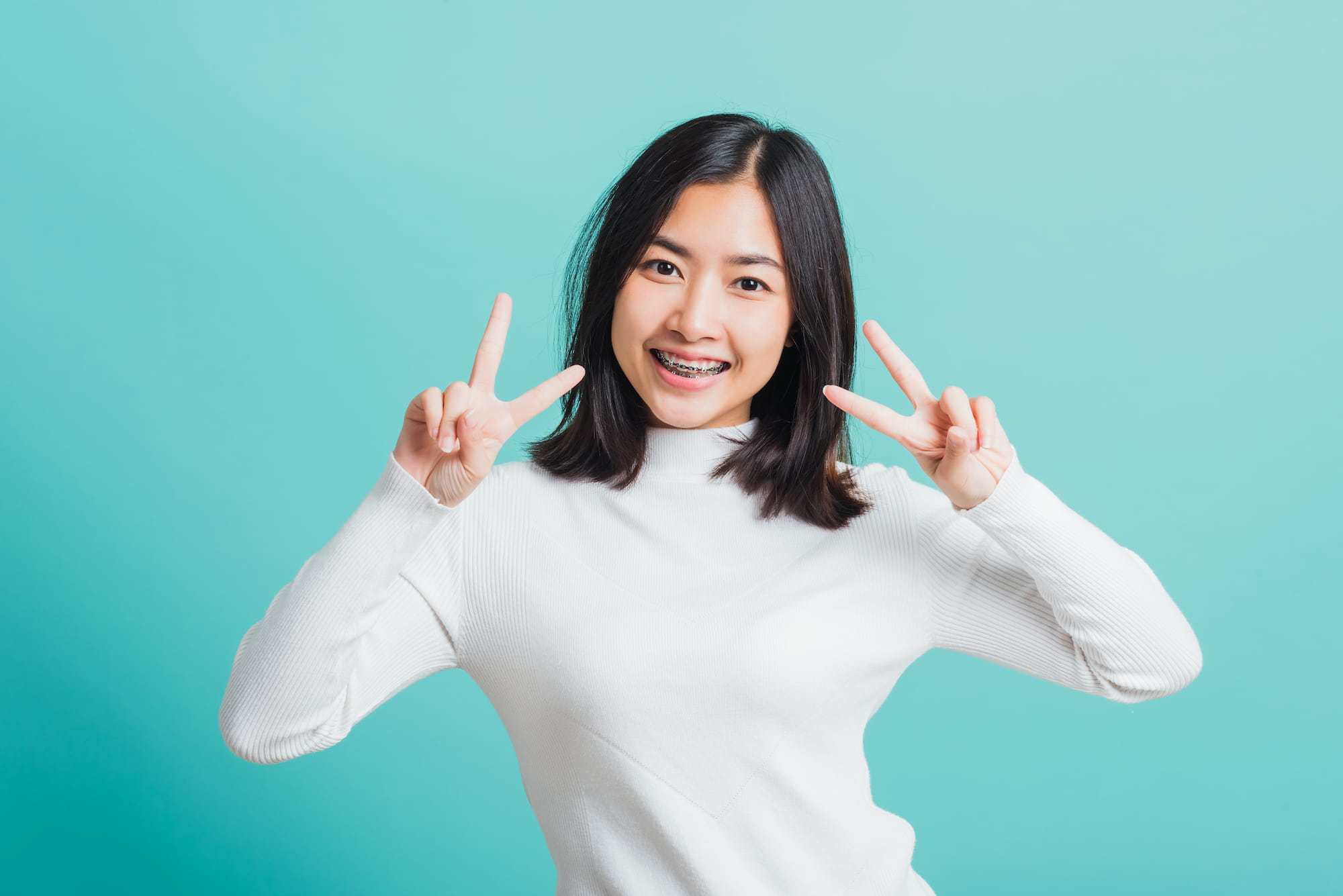 Radiating confidence after her visit to Andros Orthodontics in Pasco, WA, a young woman with shoulder-length dark hair and braces smiles and flashes double peace signs against a light blue background.