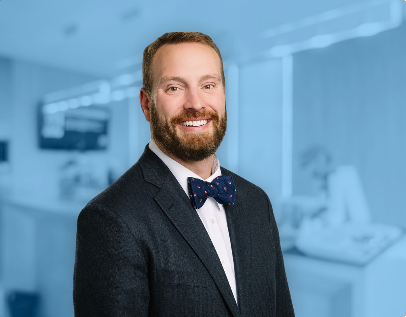 Dr. Nicholas Andros is pictured smiling at the camera in a dark suit with a white shirt and navy blue bow tie against a plain gray background. Dr. Nicholas Andros is orthodontics expert at Andros Orthodontics in Pasco, WA